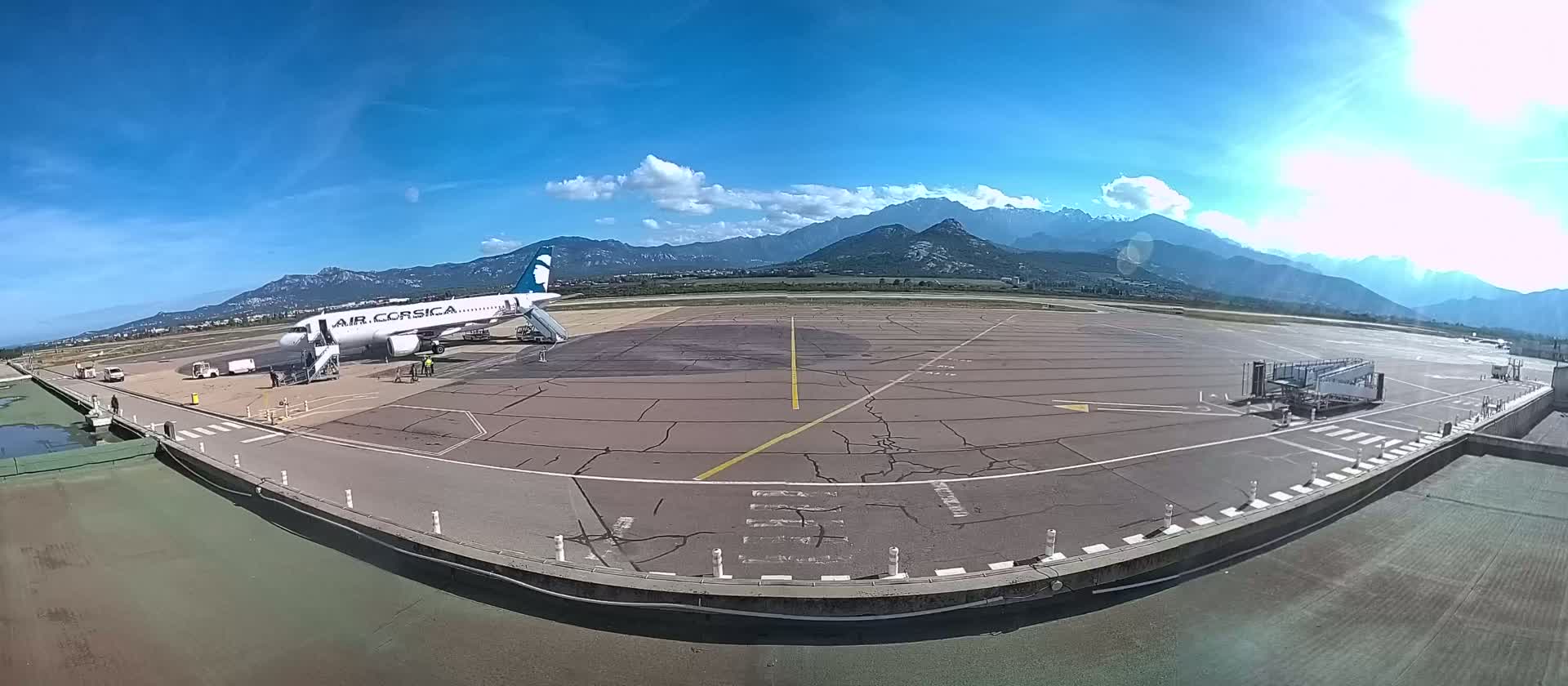 An airplane is parked at a gate on an airport tarmac under a sunny sky with mountains in the background.
