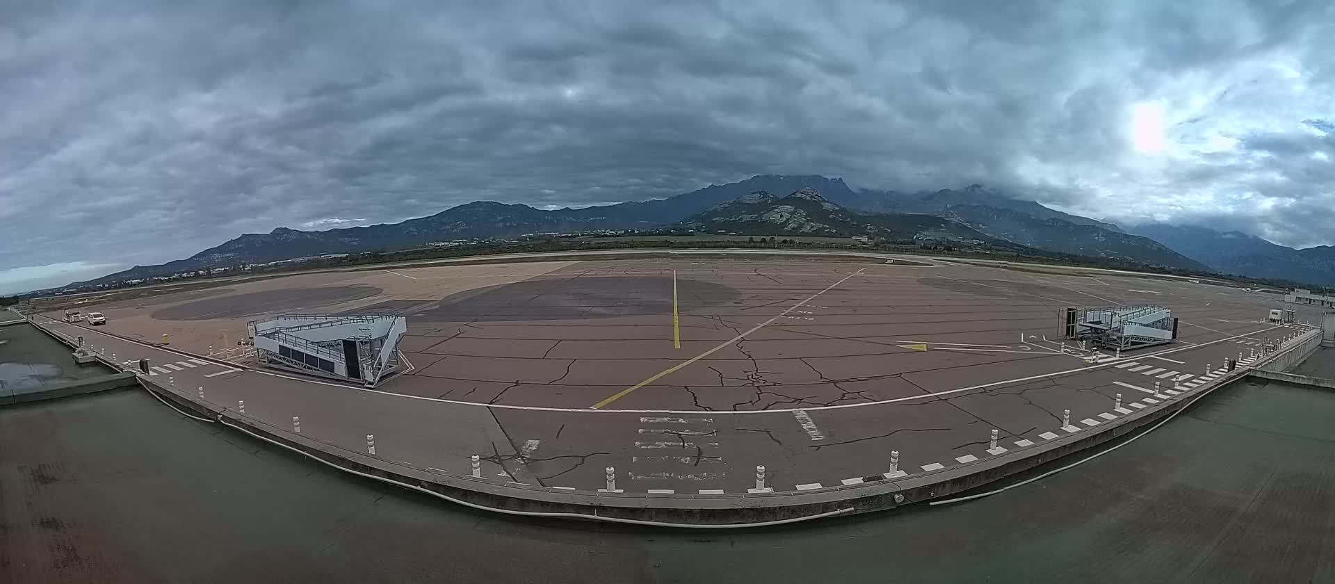 An airport tarmac with two passenger boarding bridges is visible under a cloudy sky, with mountains in the background.