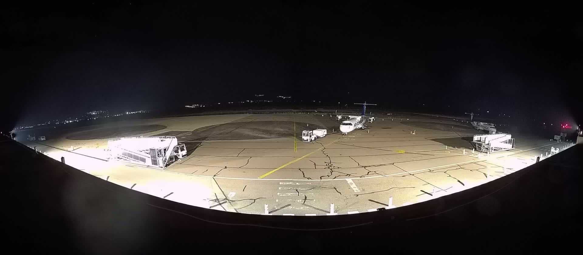 A nighttime, brightly lit view of an airport tarmac shows a small airplane parked between two jet bridges and several service vehicles.