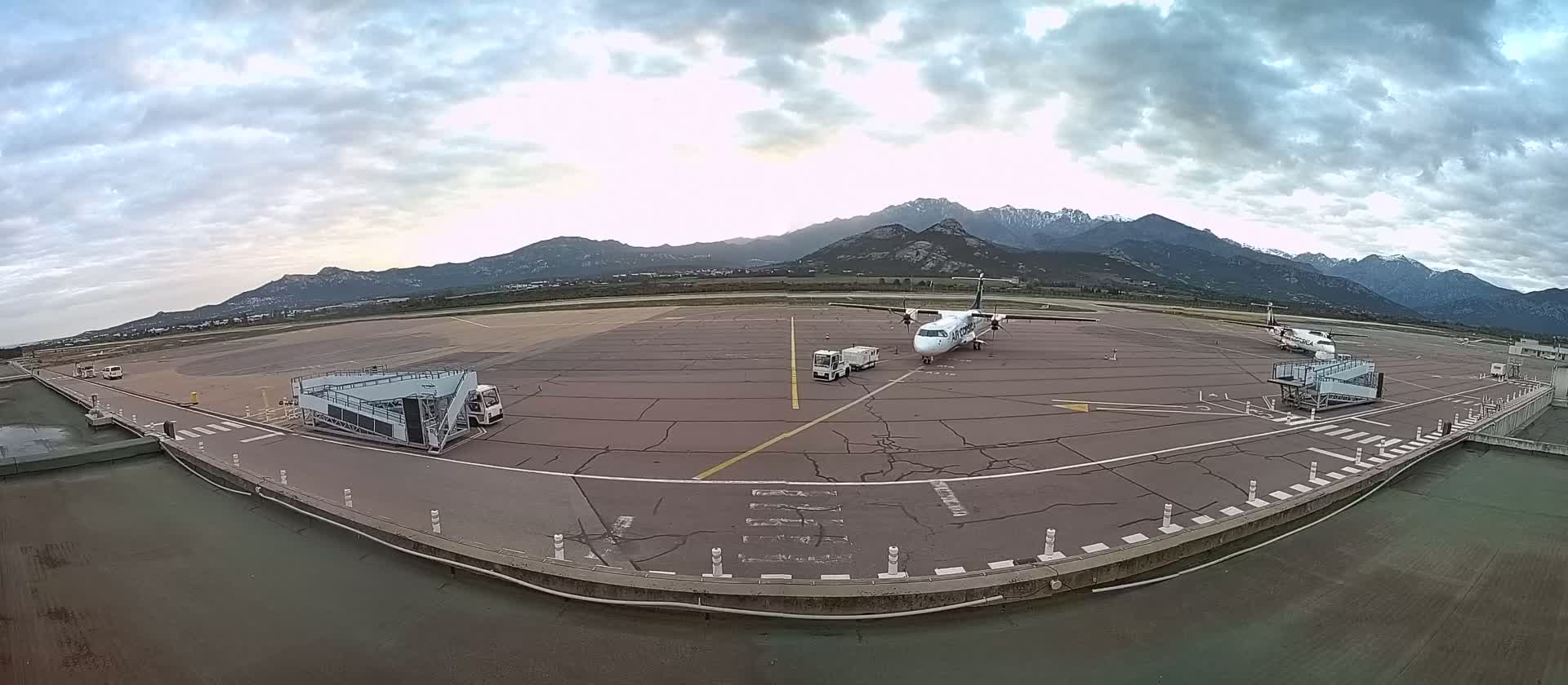 A small airplane is parked on an airport tarmac under a cloudy sky, with mountains visible in the background.