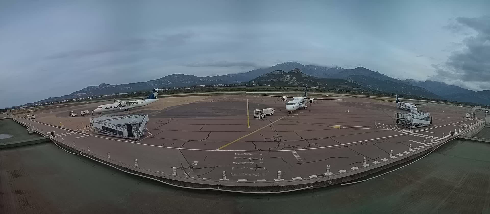 Two airplanes are parked at a tarmac with passenger boarding bridges, service vehicles nearby, and mountains in the background under an overcast sky.