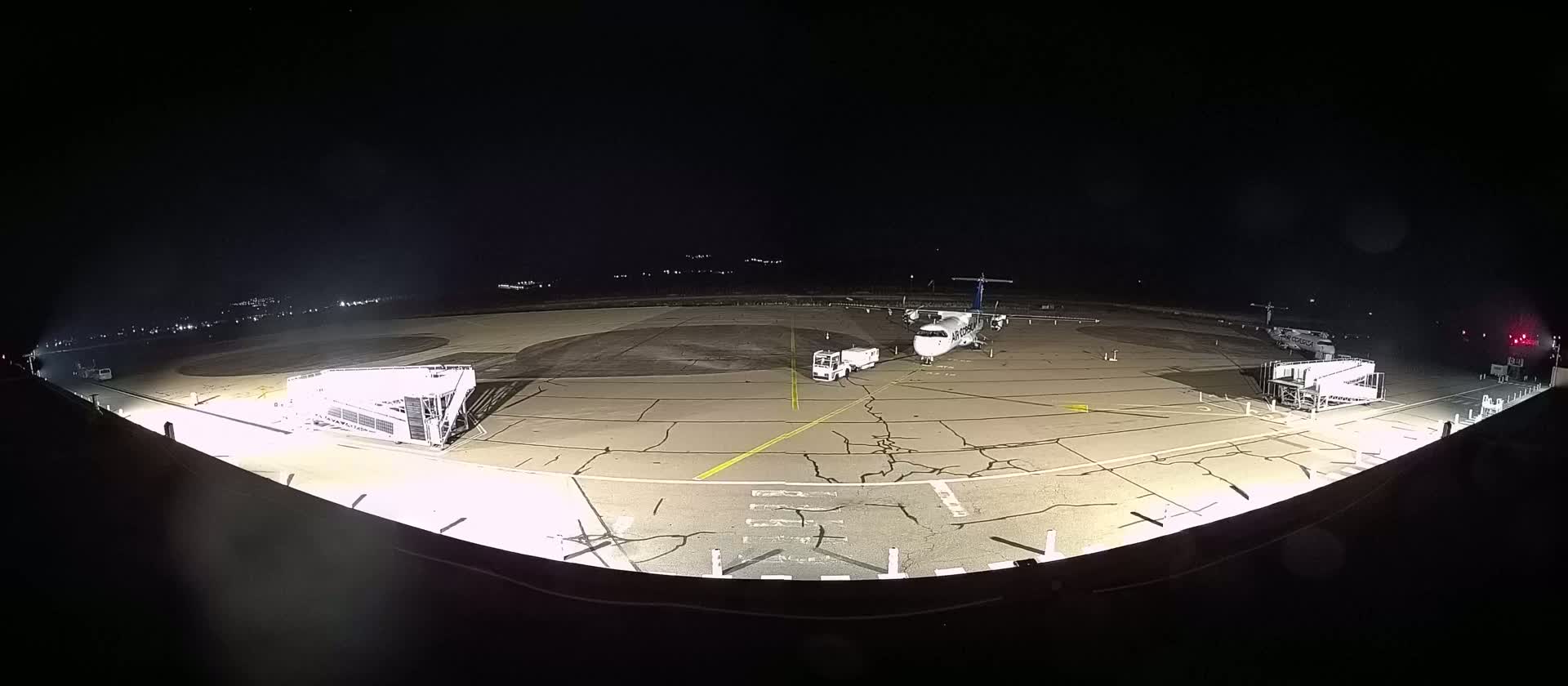 A propeller plane sits on a tarmac at night, flanked by two jet bridges and a service vehicle, under a clear, dark sky.