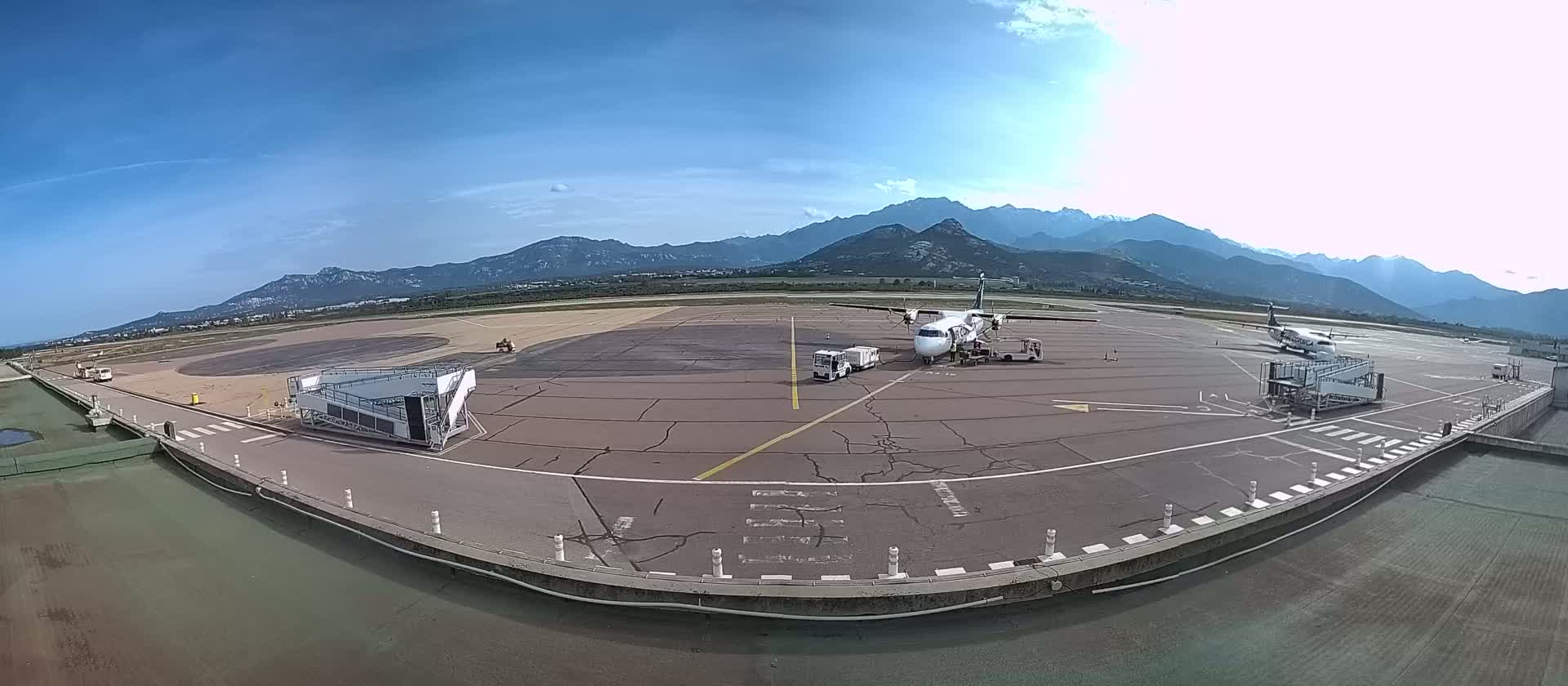 A turboprop airplane is parked at a gate on an airport tarmac with two jet bridges and service vehicles present, under a clear, sunny sky with mountains in the background.