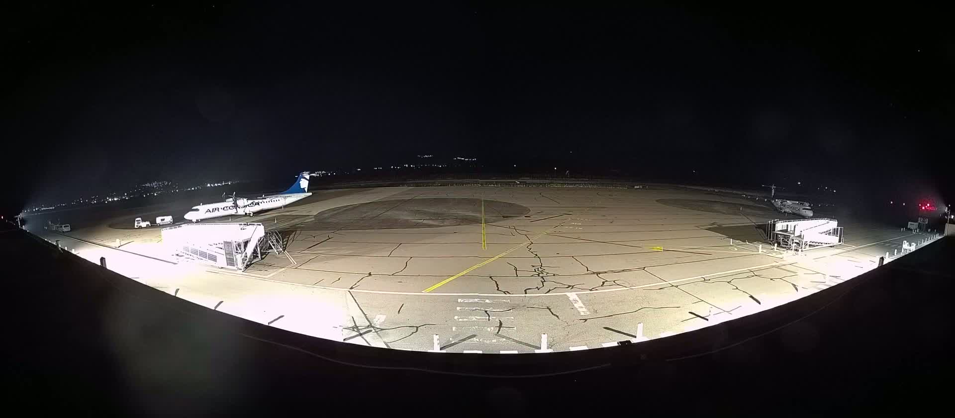 A nighttime, brightly lit airport tarmac shows a passenger plane connected to a jet bridge, with another plane nearby and several ground vehicles visible.