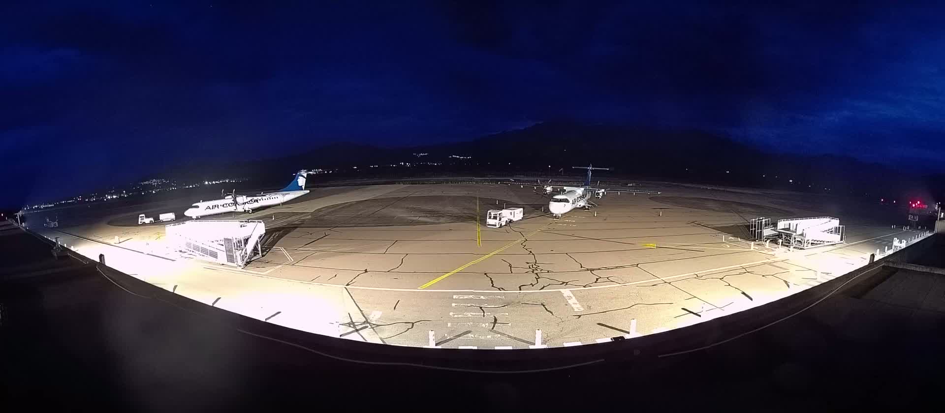 Two airplanes are parked at night on a tarmac under a dark blue sky, illuminated by bright lights, with ground support equipment nearby.