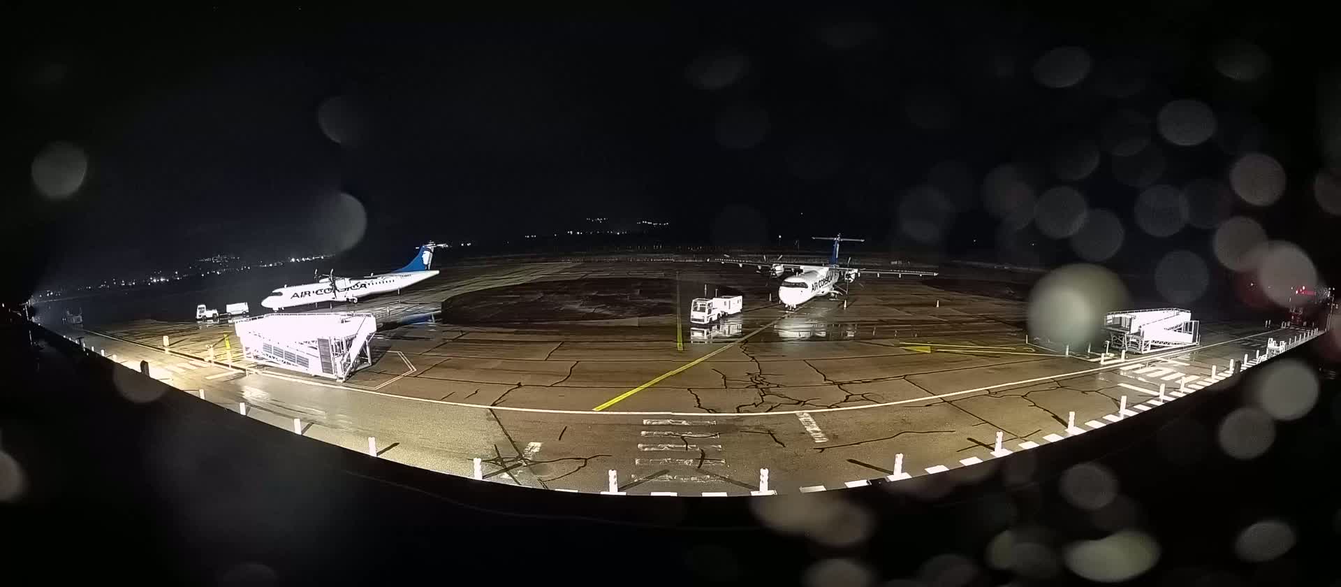 Two airplanes are parked on a rain-slicked tarmac at night, with ground support equipment nearby.