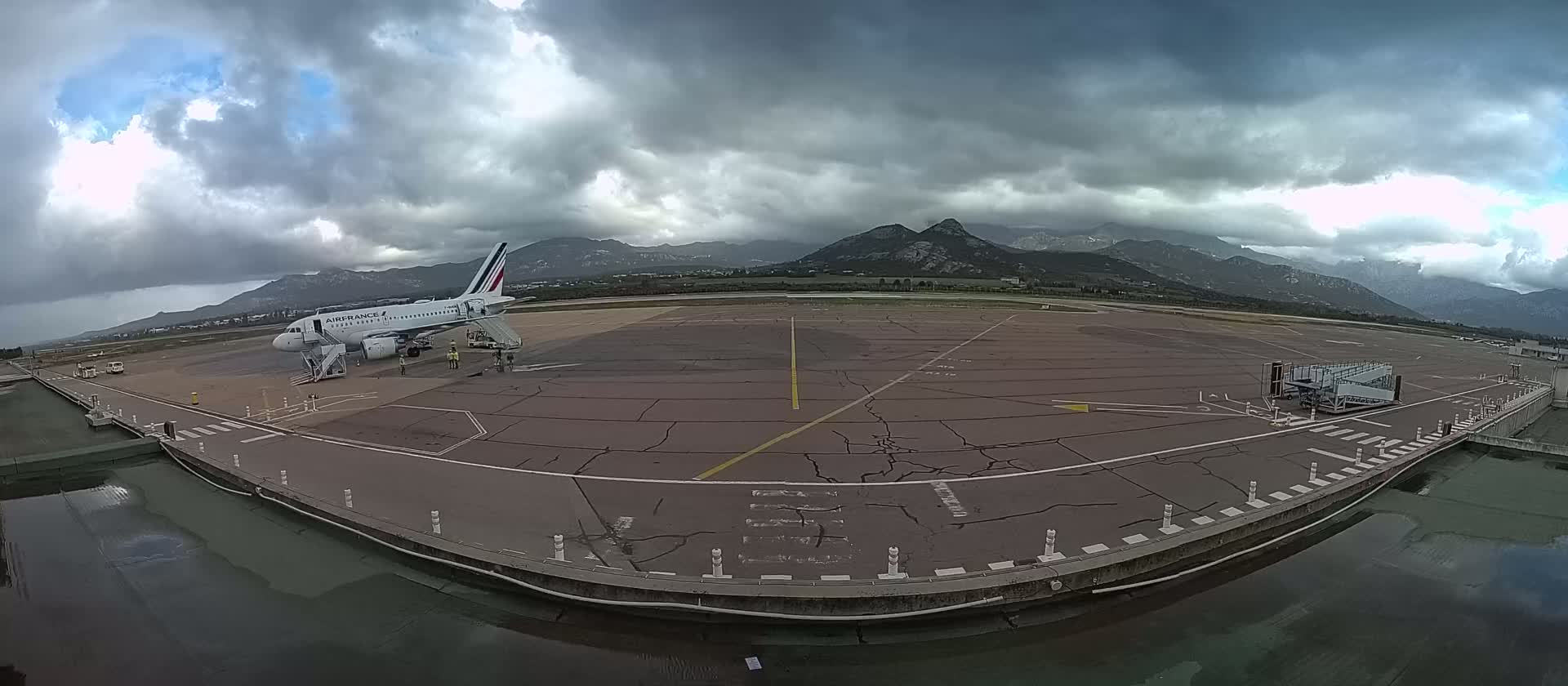 An airplane is parked at a tarmac next to a jet bridge, with overcast skies and mountains in the background.