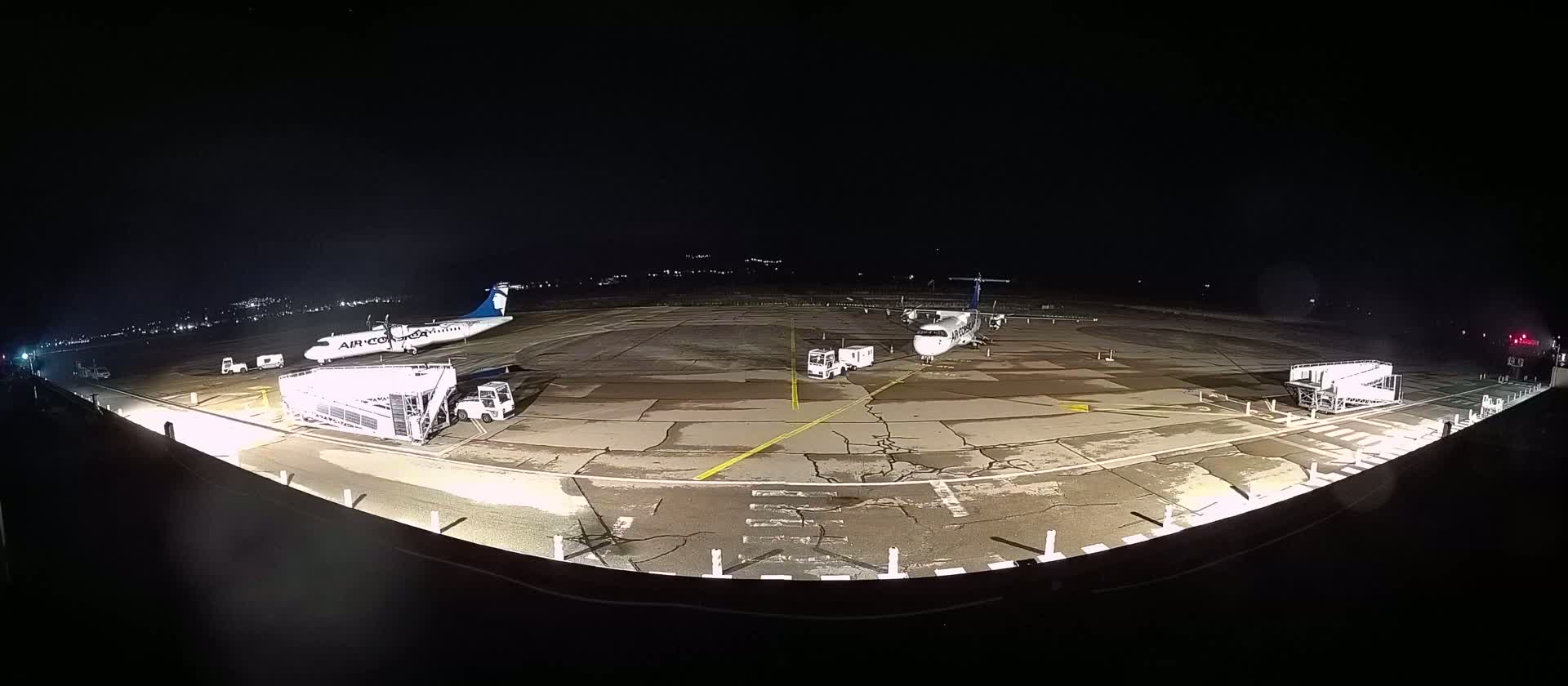 At night, two airplanes are parked on a tarmac with ground support vehicles and jet bridges.