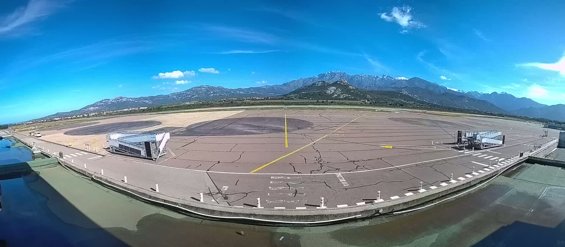 An empty airport tarmac with two jet bridges is seen under a clear blue sky with mountains in the distance.