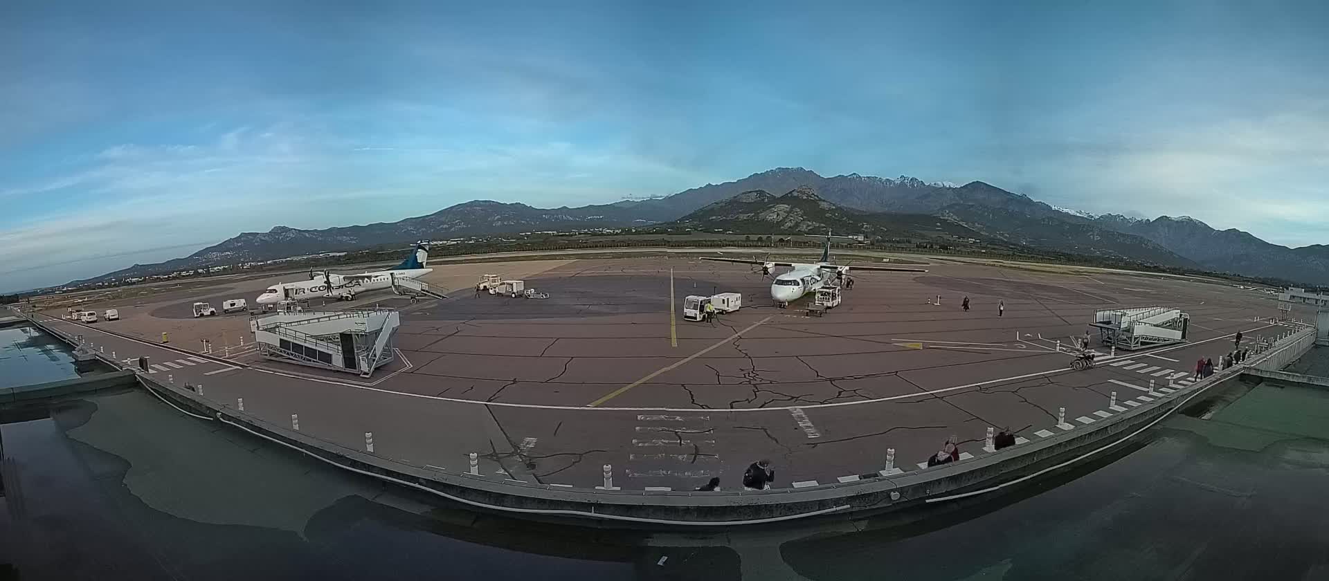 Two airplanes are parked on an airport tarmac under a clear sky, with mountains visible in the distance.