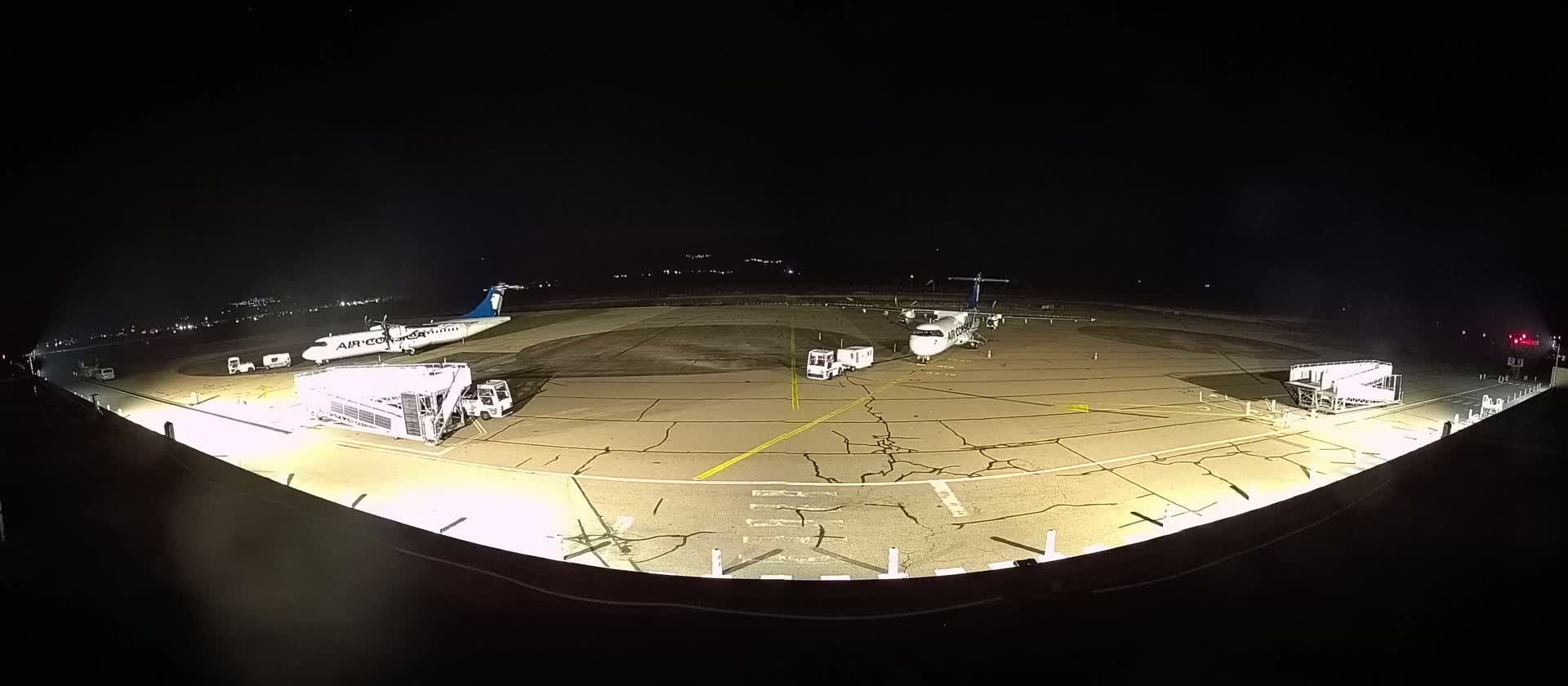 At night, two airplanes are parked on a tarmac with ground support equipment and jet bridges.
