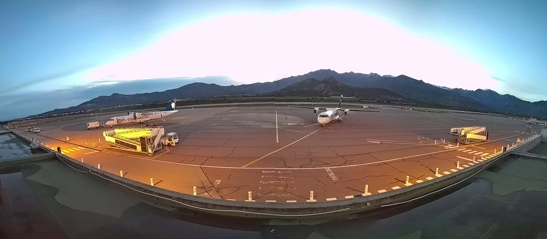 Two airplanes are parked on an airport tarmac at dusk, with mountains in the background.