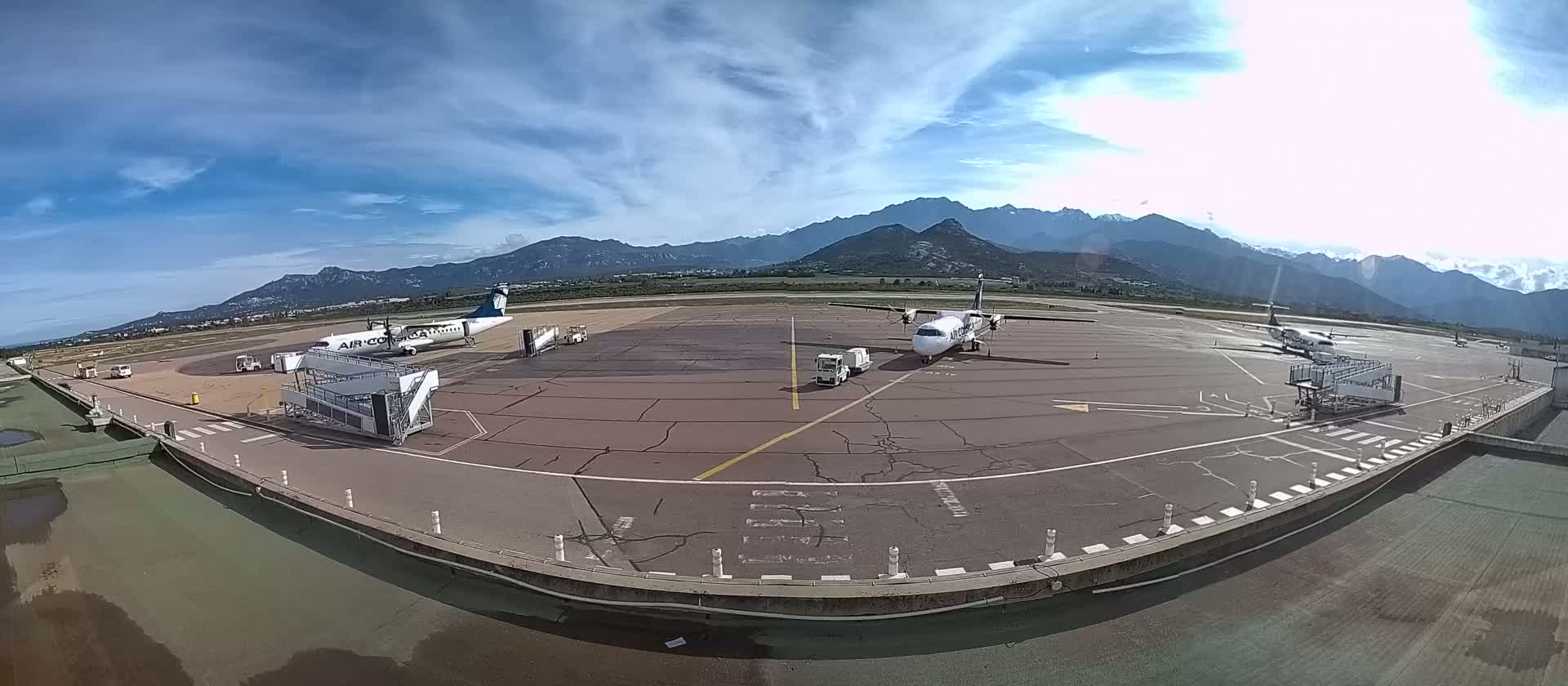 Three airplanes are parked at a tarmac next to jet bridges, with mountains visible in the background under a partly cloudy sky.