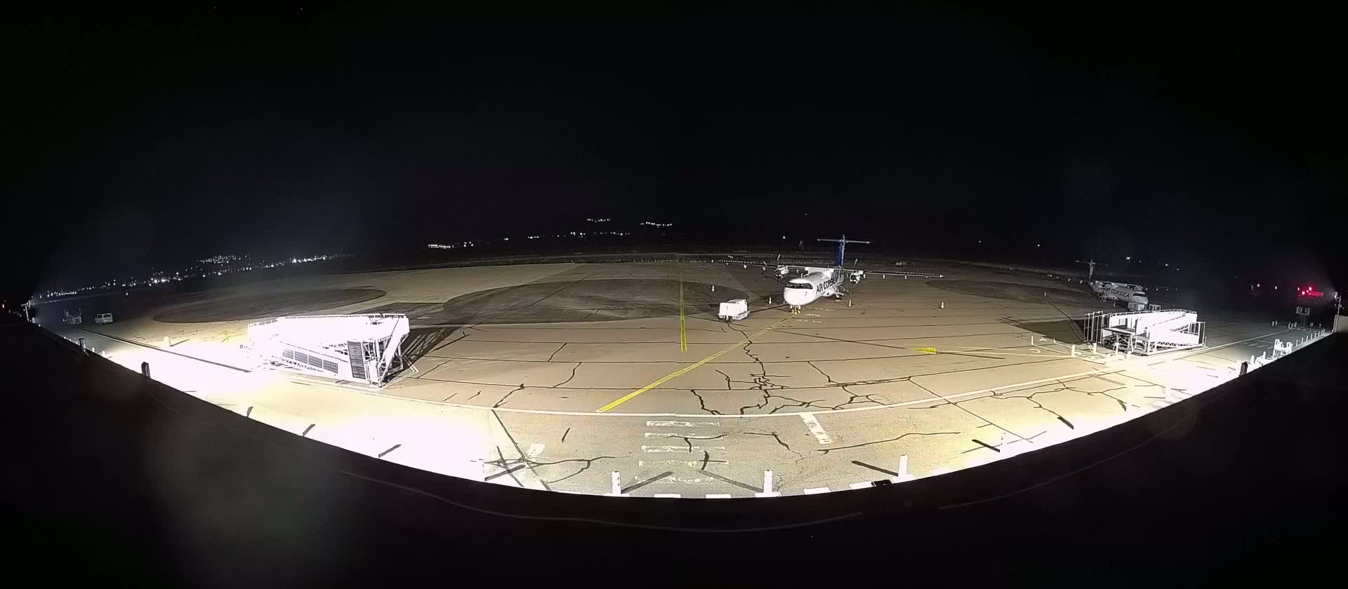 At night, a small airplane sits on a tarmac next to two passenger boarding bridges, surrounded by cracked pavement and sparsely lit by floodlights.