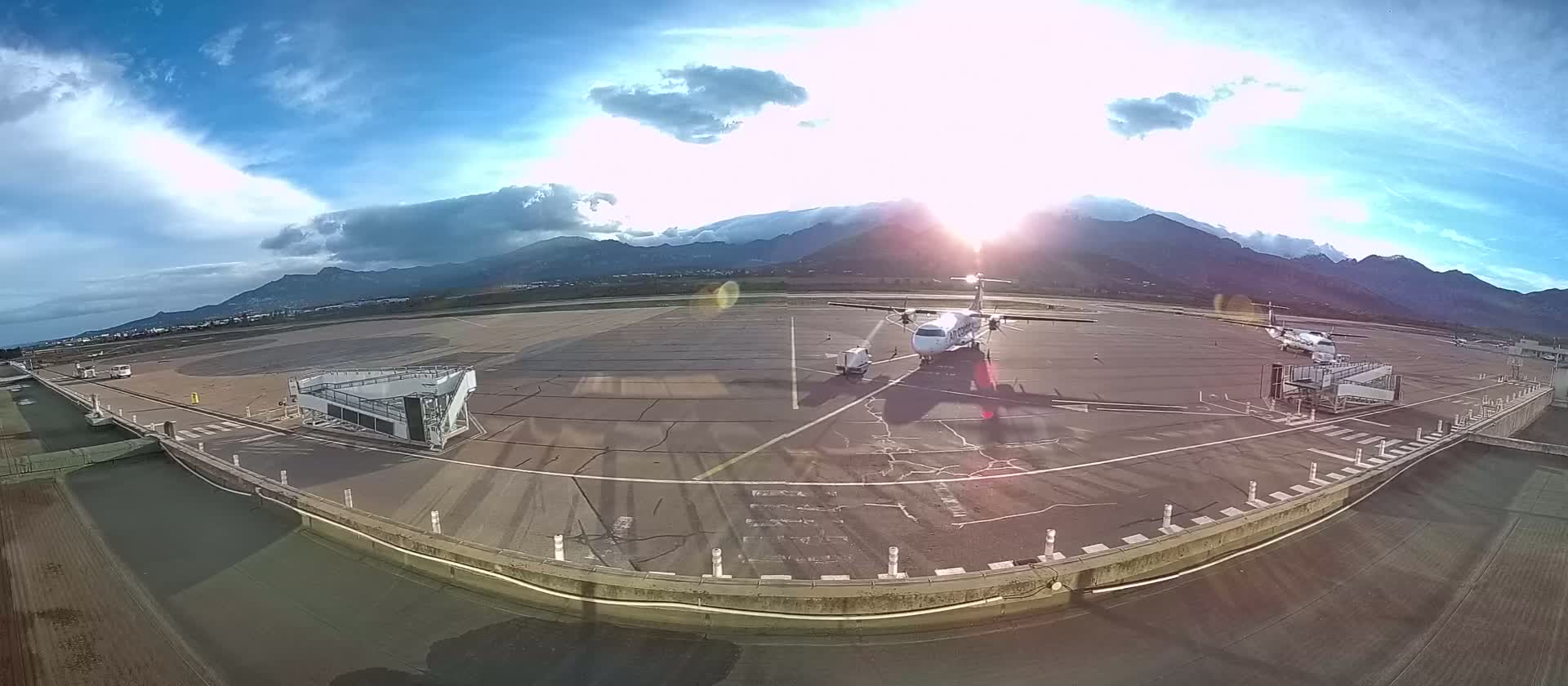 A propeller plane sits on a tarmac at an airport against a backdrop of mountains under a partly sunny sky.