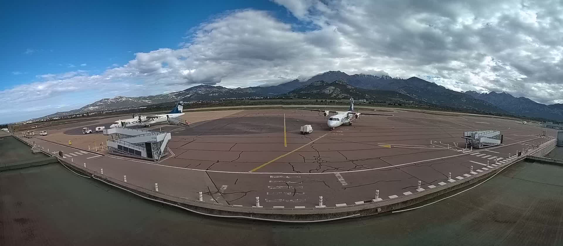 Two airplanes are parked on an airport tarmac under a partly cloudy sky, with mountains visible in the background.