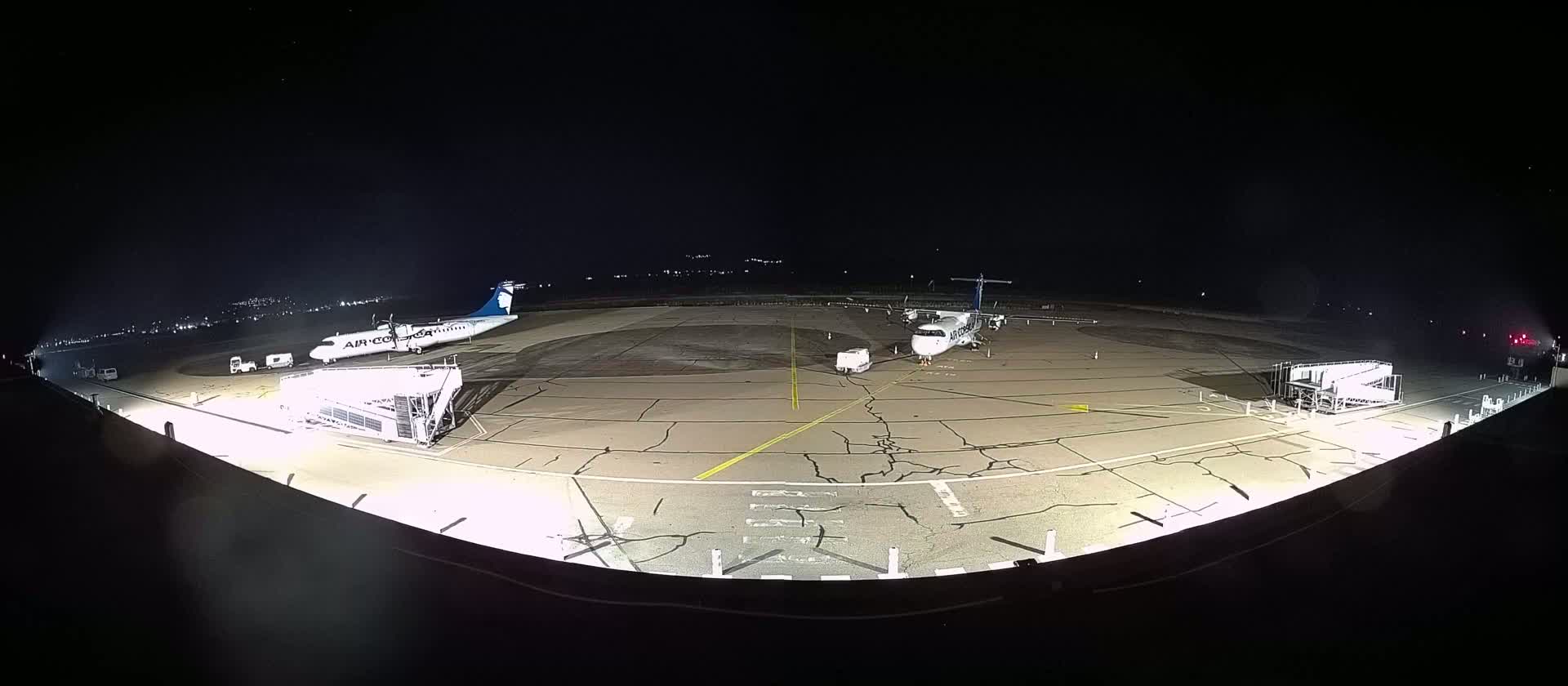 At night, two airplanes are parked on a tarmac, one with a jet bridge attached, illuminated by bright lights.