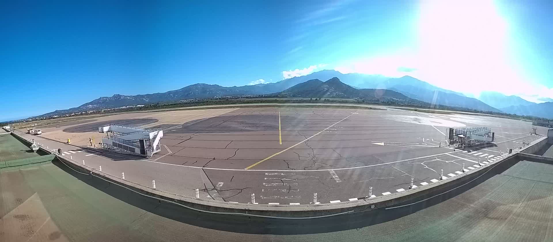 A tarmac at an airport with two passenger boarding bridges, under sunny skies with mountains in the background.