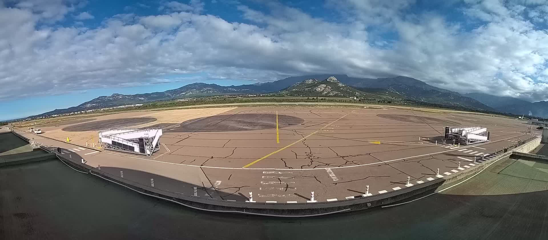 An airport tarmac with two jet bridges is visible, under a partly cloudy sky, with mountains in the background.