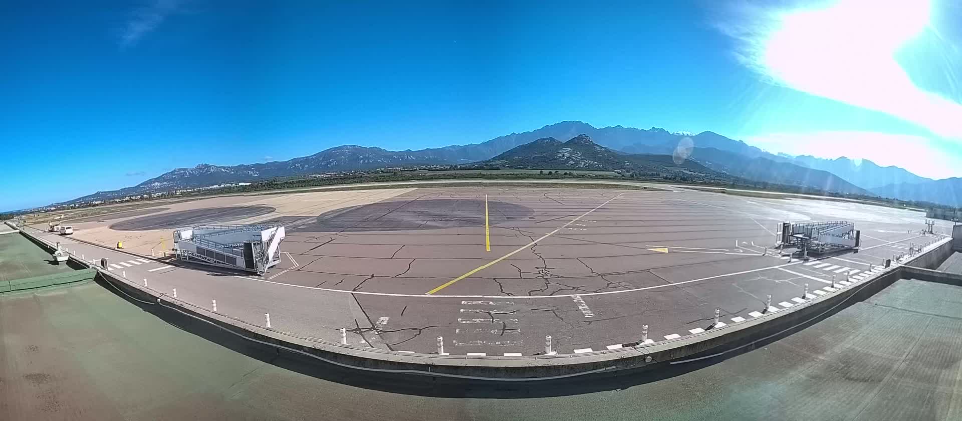 An airport tarmac with two passenger boarding bridges is visible under a clear, sunny sky with mountains in the background.