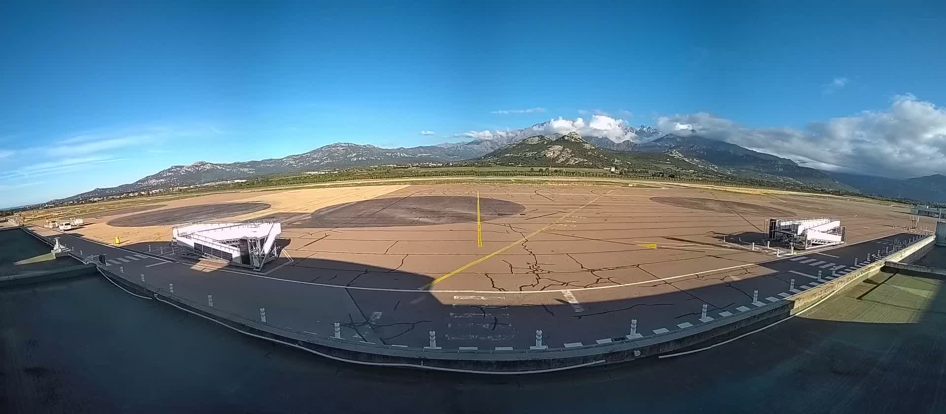 An airport tarmac with two jet bridges is shown under a clear blue sky, with mountains in the background partially obscured by clouds.