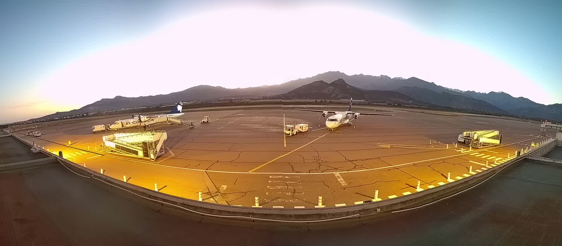 Two airplanes are parked on an airport tarmac at sunrise, with mountains visible in the background.