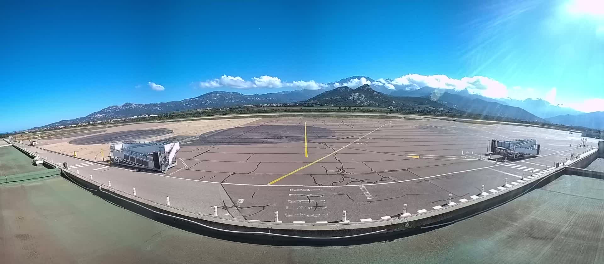 An airport tarmac with two passenger boarding bridges, under a sunny sky and backed by a mountain range.