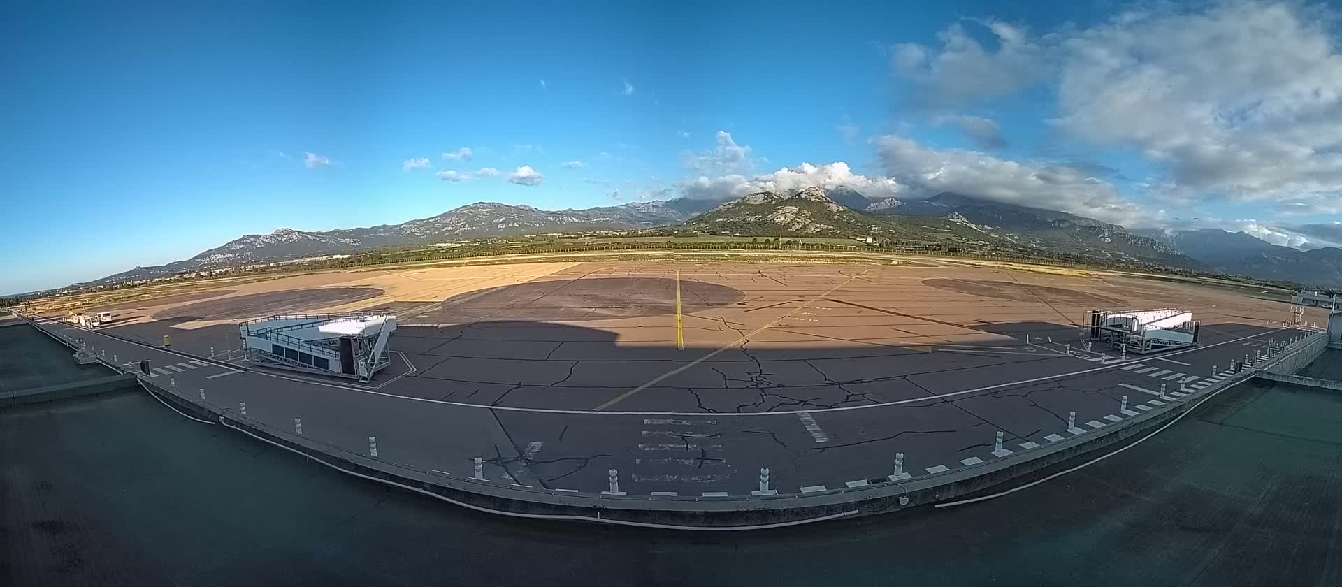 An airport tarmac with two jet bridges is visible, under a partly cloudy blue sky, with mountains in the background.