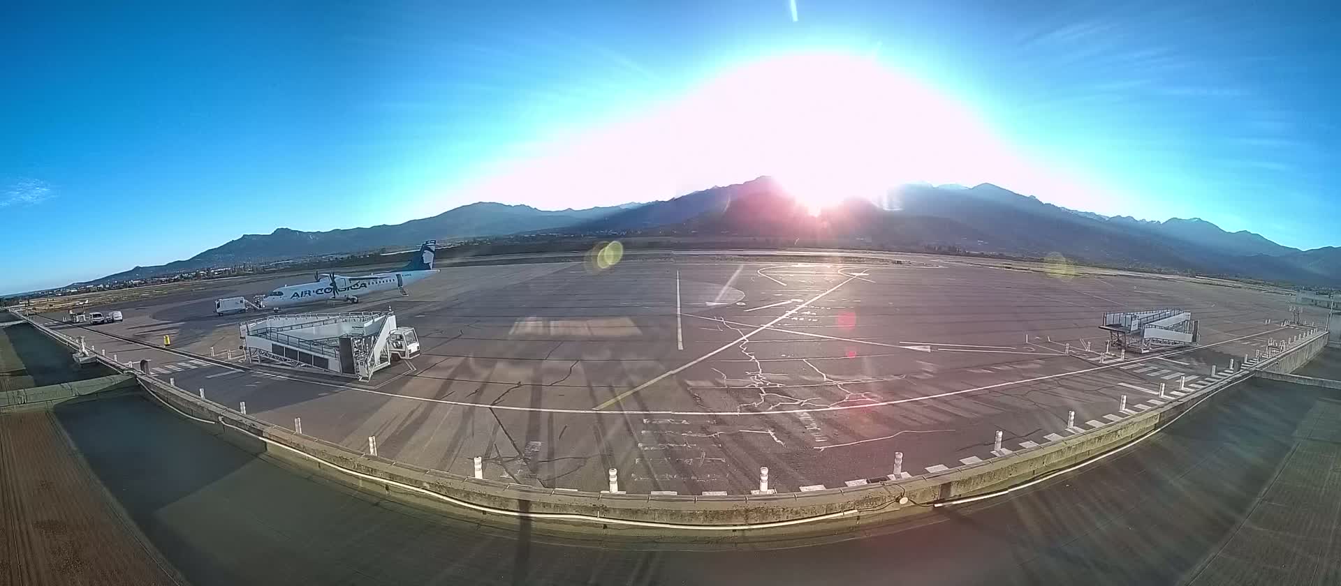 A plane sits at a gate on a sunny airport tarmac, backed by a mountain range under a clear blue sky.