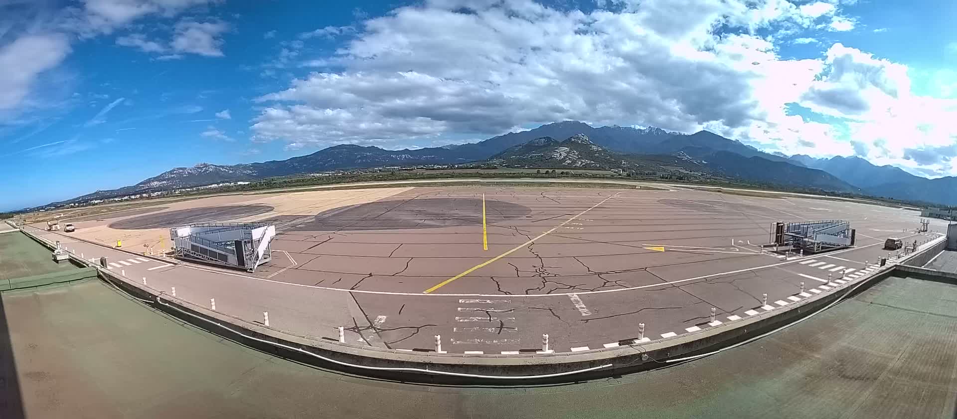 An airport tarmac with two passenger boarding bridges is visible, under a partly cloudy sky with mountains in the background.