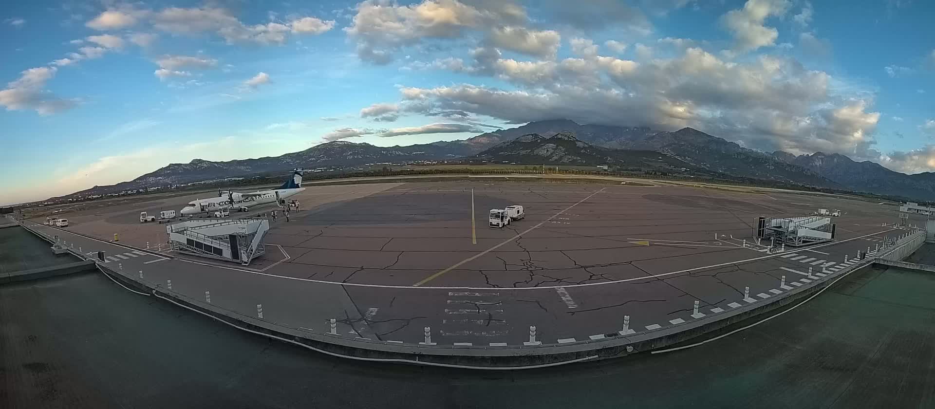 A partly cloudy sky overlooks an airport tarmac with a single airplane parked at a jet bridge, service vehicles, and mountains in the distance.