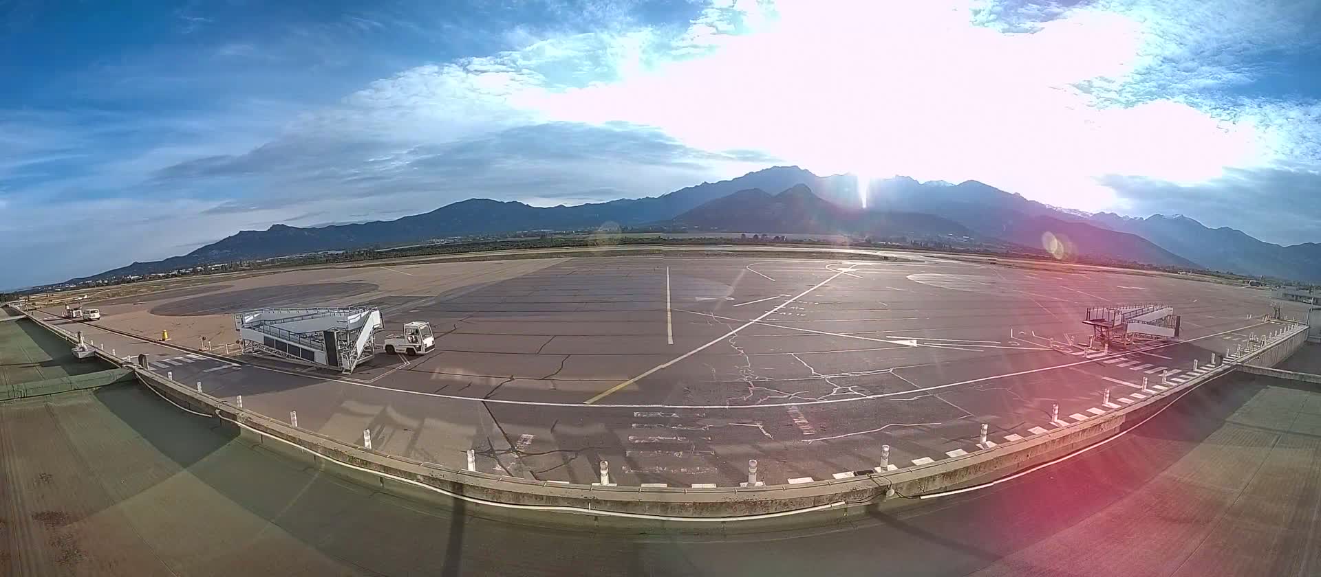 An airport tarmac with a jet bridge and support vehicle is shown under a partly cloudy sky, with mountains visible in the distance.