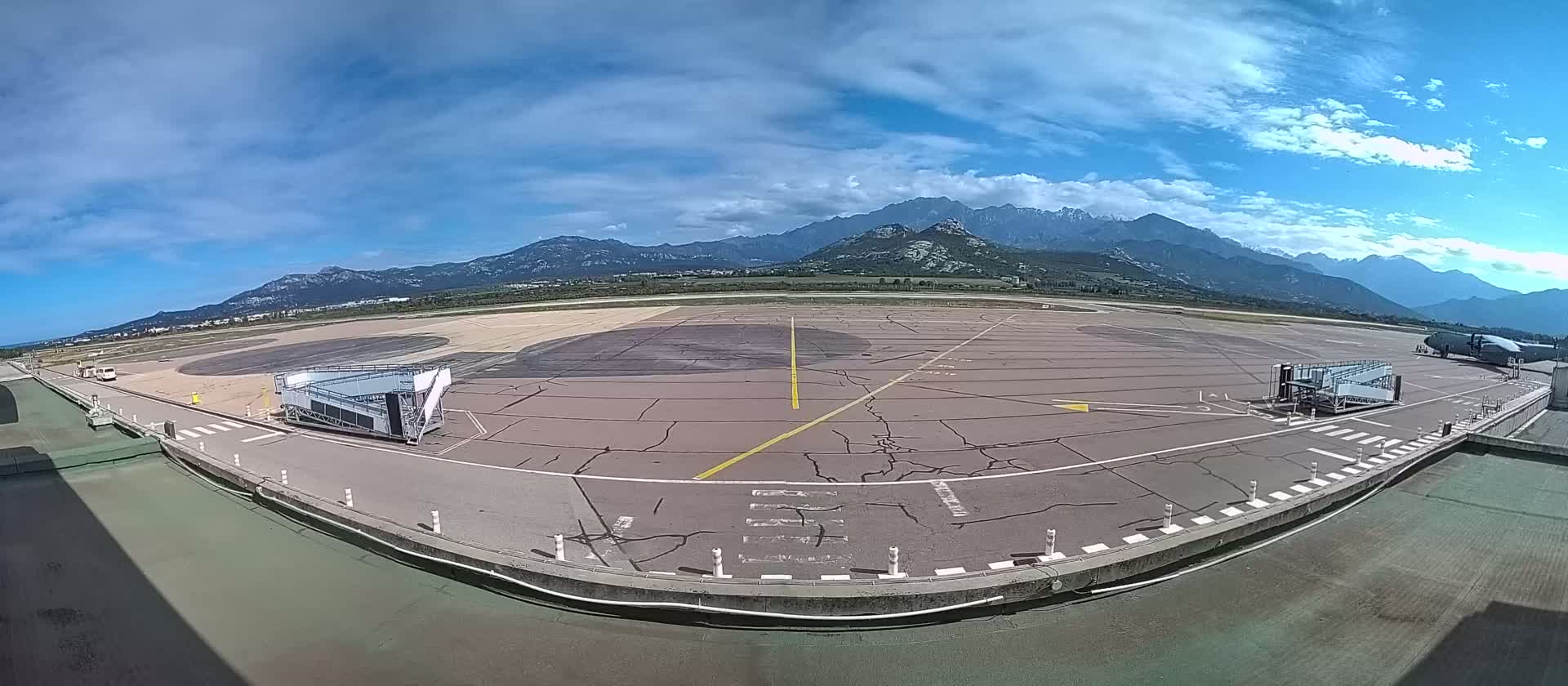 An airport tarmac with two jet bridges, under a partly cloudy sky, is situated against a backdrop of mountains.