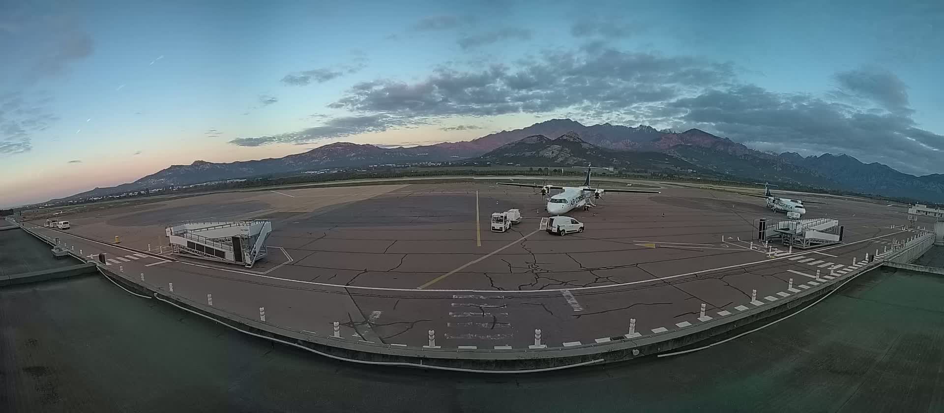 Two airplanes are parked on an airport tarmac at dusk, with mountains visible in the background under a partly cloudy sky.