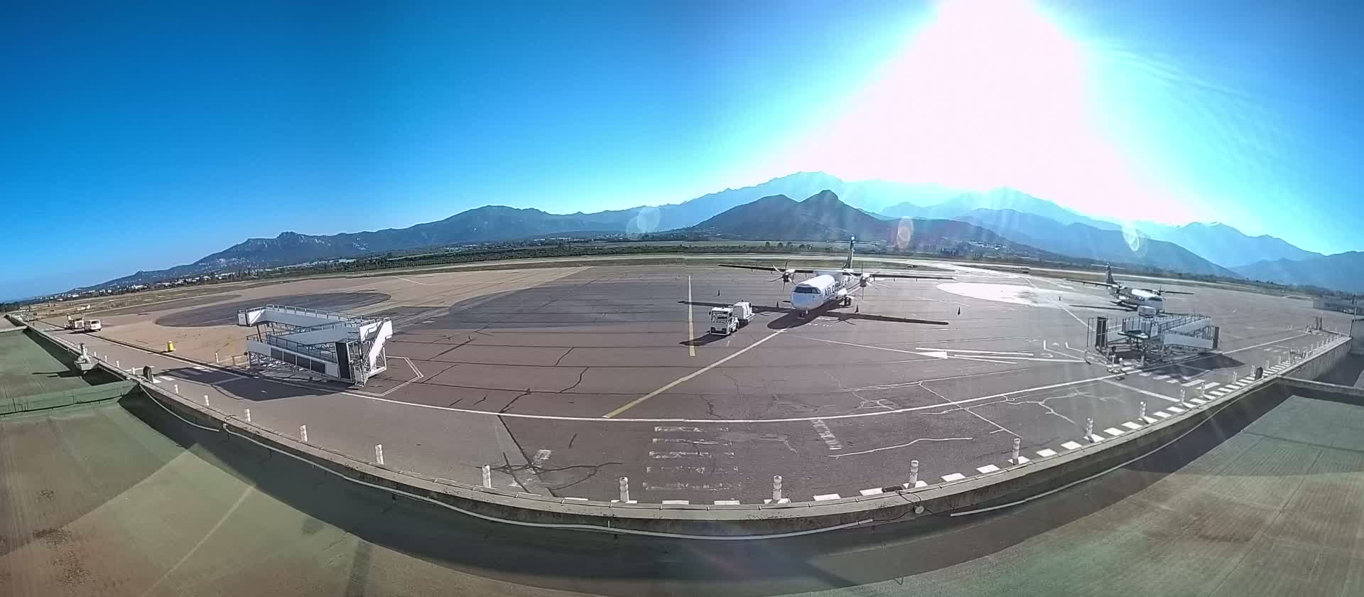 Two airplanes are parked at a tarmac on a sunny day with mountains in the background.