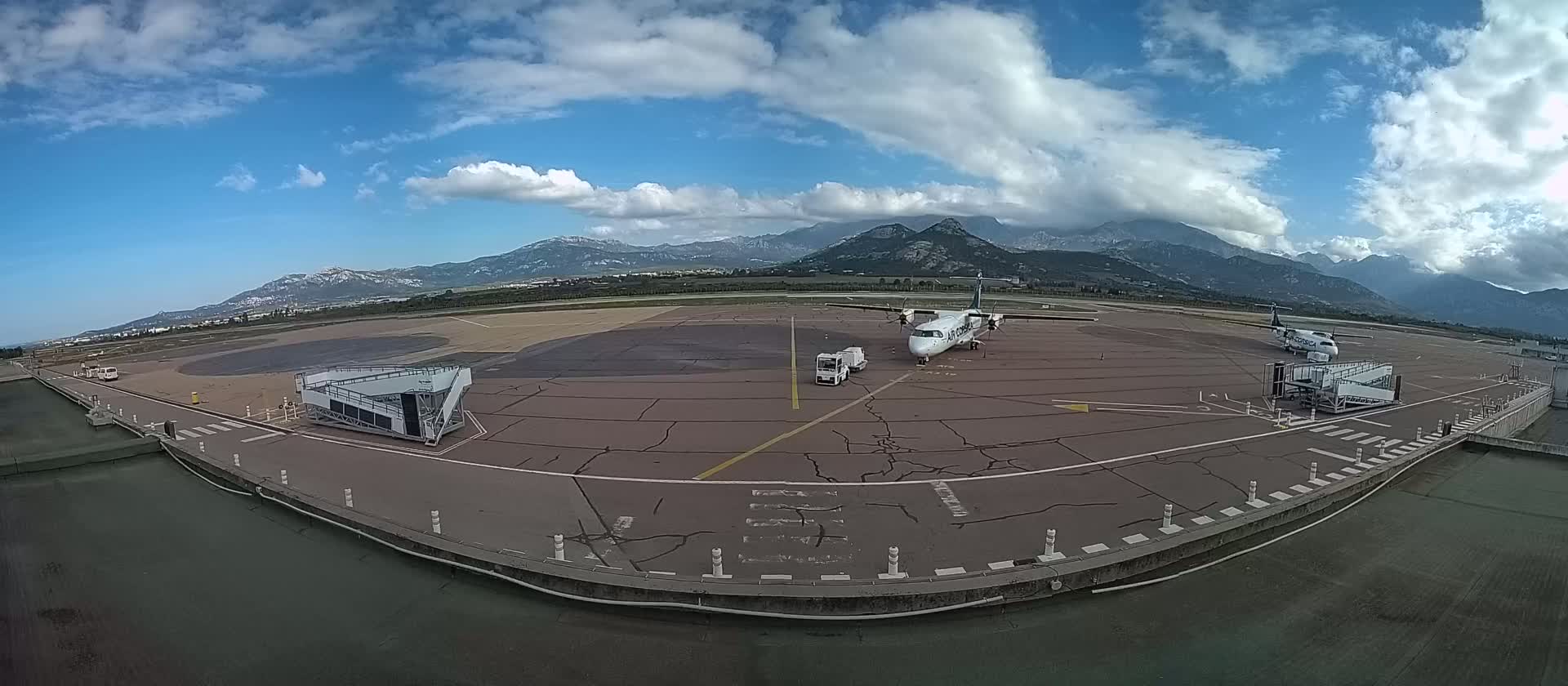 A tarmac view shows two airplanes parked at gates with jet bridges, service vehicles, and a mountainous backdrop under a partly cloudy sky.