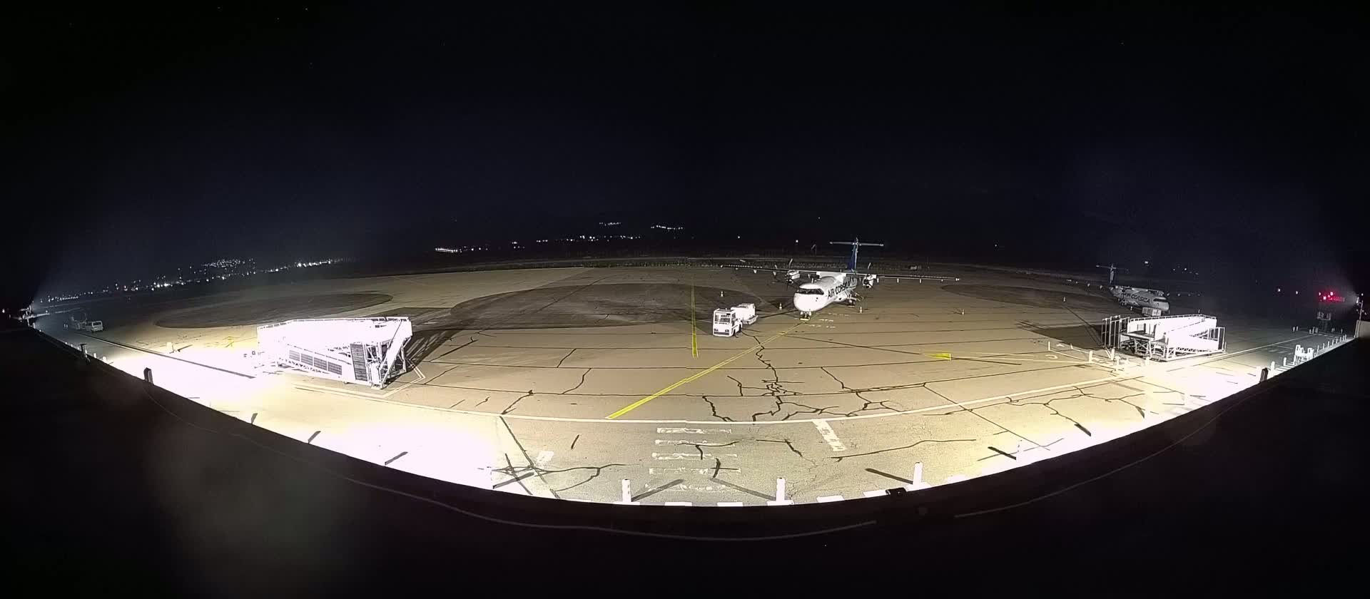 A turboprop airplane is parked on a tarmac at night, illuminated by lights, with jet bridges and service vehicles nearby.