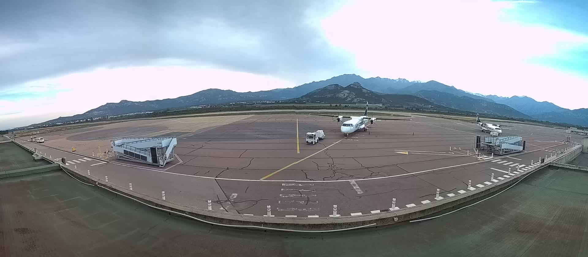 An airport tarmac with two airplanes parked near jet bridges, backed by a mountain range under a cloudy sky.