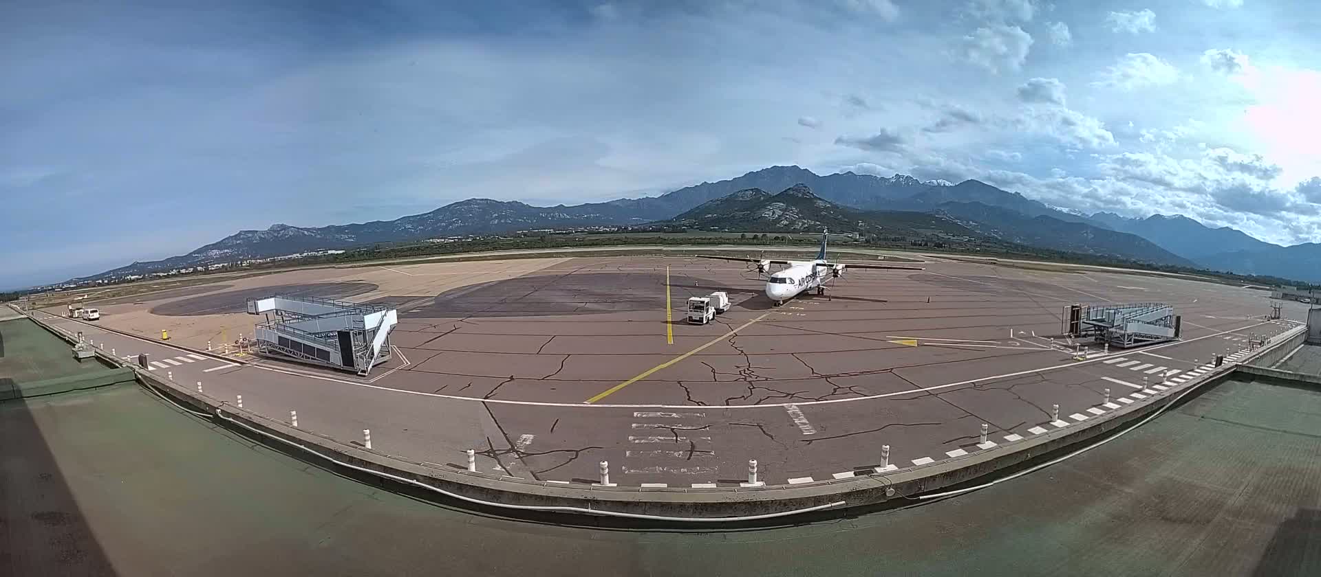 A turboprop airplane is parked at a tarmac next to two jet bridges, with mountains in the background under a partly cloudy sky.
