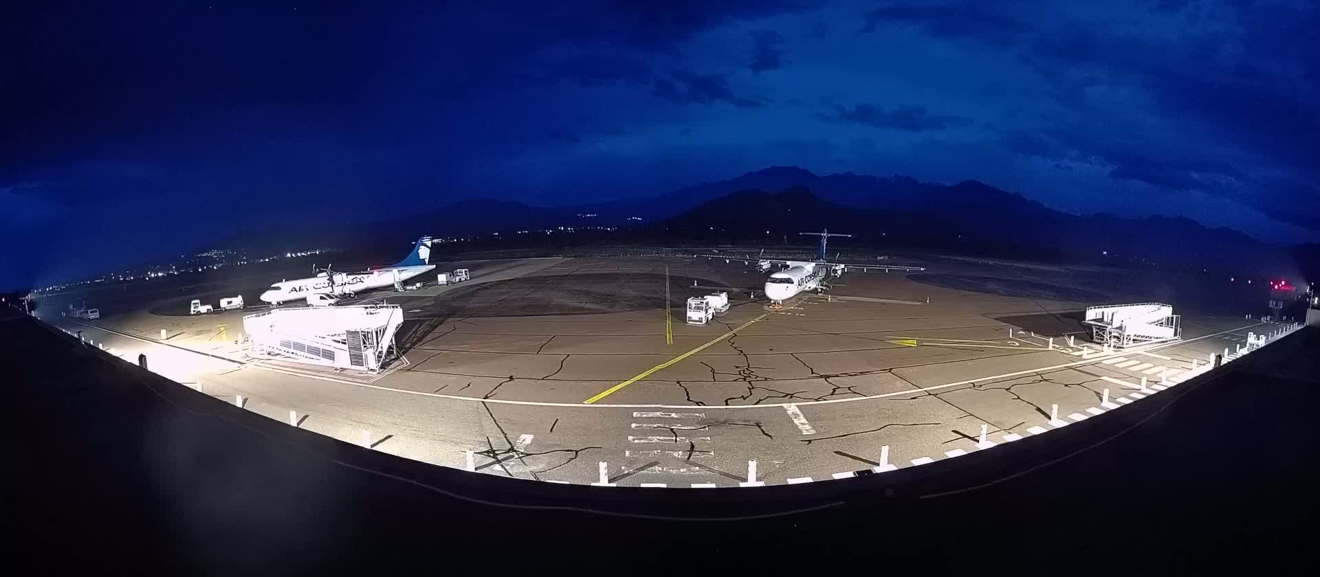 Two airplanes are parked at a nighttime airport under a dark, cloudy sky with mountains in the distance.