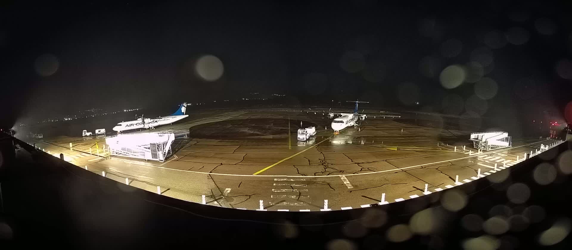 Two airplanes are parked at night on a rain-slicked tarmac, with jet bridges and support vehicles nearby.