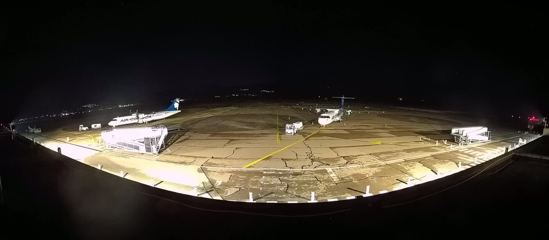 At night, two airplanes are parked on a tarmac illuminated by bright lights, with ground support equipment nearby.