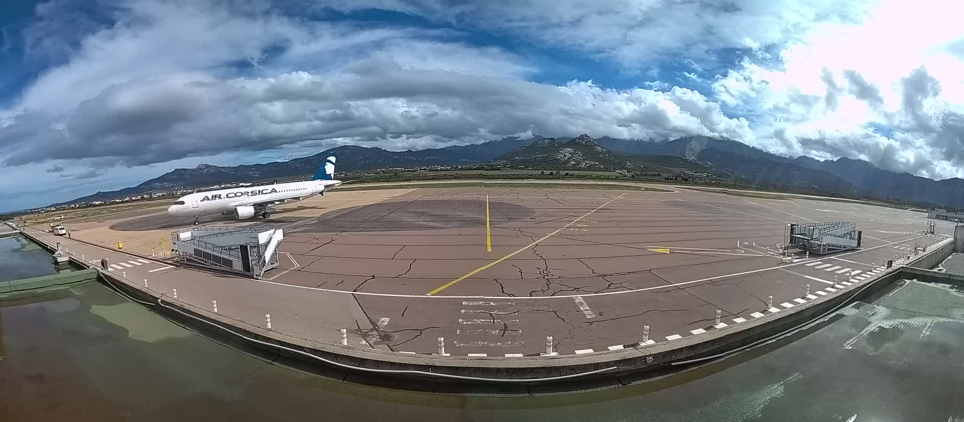 An airplane is parked at a gate on an airport tarmac under a partly cloudy sky, with mountains visible in the background.