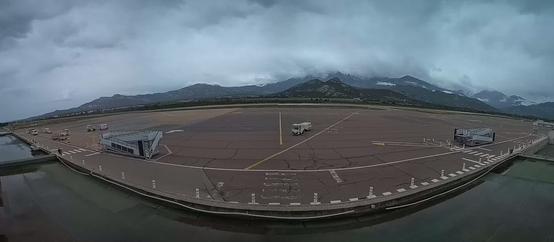 An empty airport tarmac with two jet bridges and a service vehicle, under an overcast sky with mountains in the background.