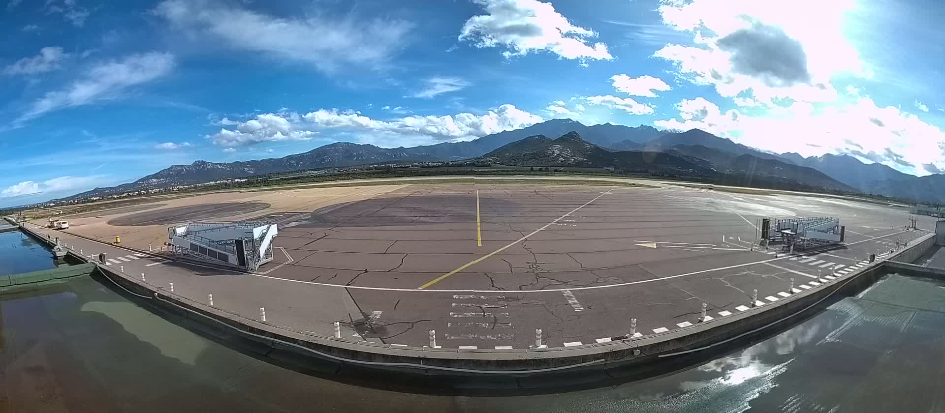 A mostly sunny day at an airport tarmac with a jet bridge, featuring mountains in the background.
