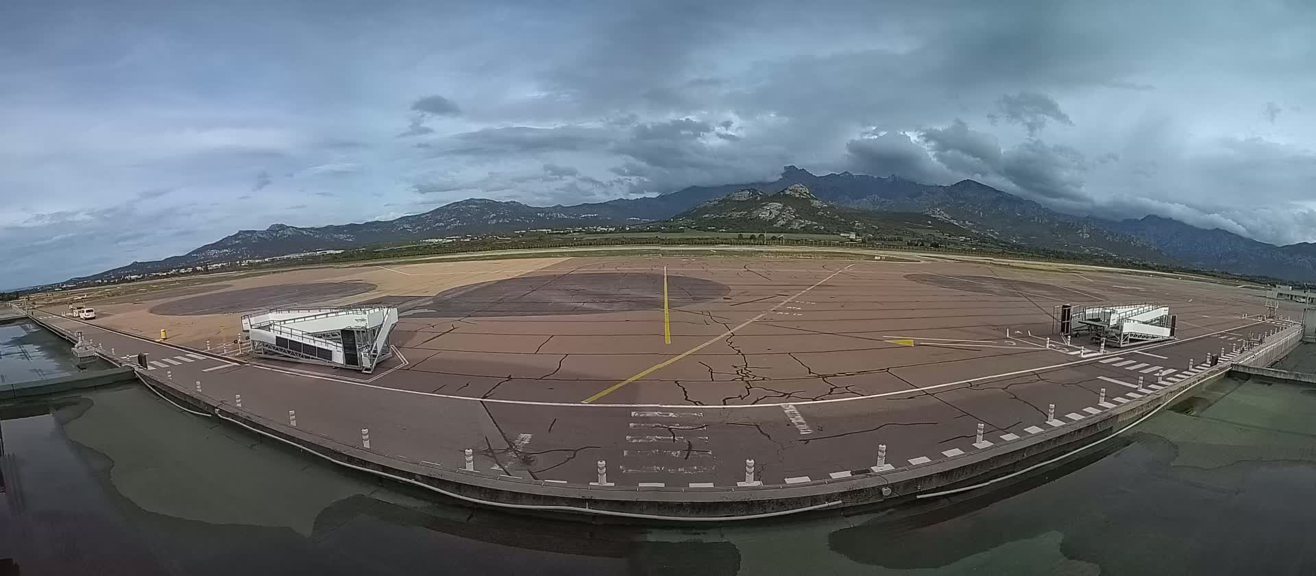 An empty airport tarmac with two passenger boarding bridges is visible under a cloudy sky, with mountains in the background.