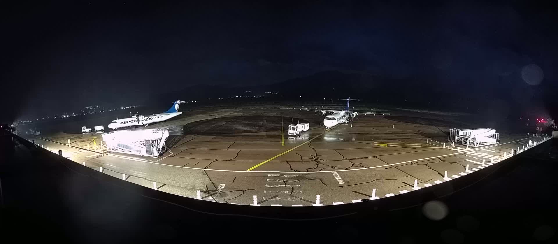 Two airplanes are parked on a wet tarmac at night, with ground support equipment nearby.