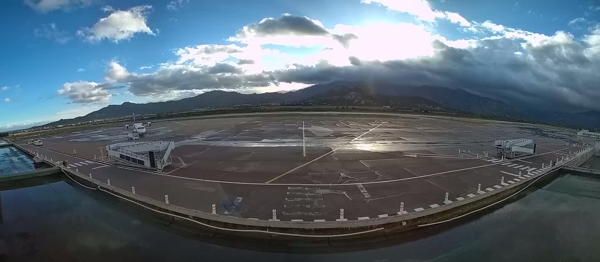 A wet airport tarmac with a plane and jet bridge is visible under a partly cloudy sky with mountains in the background.