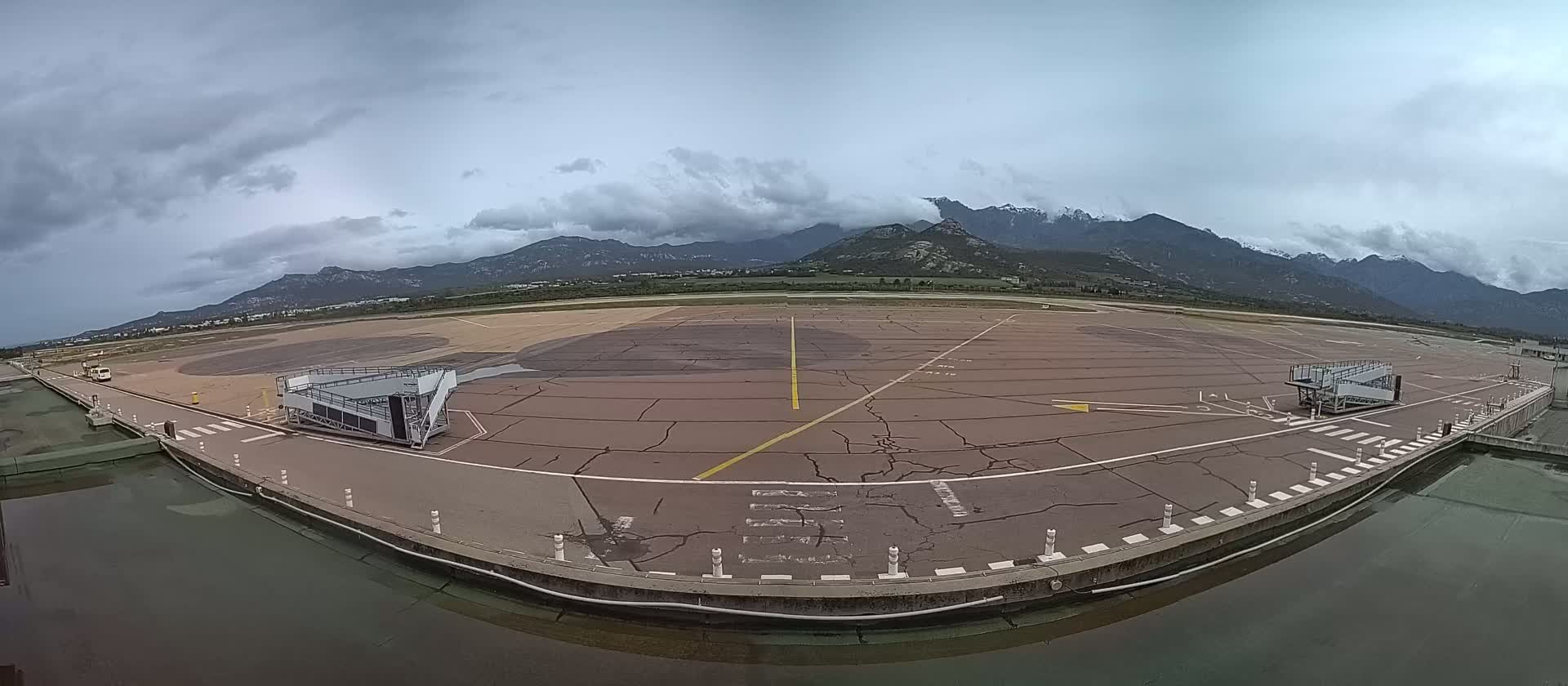 An empty airport tarmac with two passenger boarding bridges is visible under a cloudy sky, with mountains in the background.