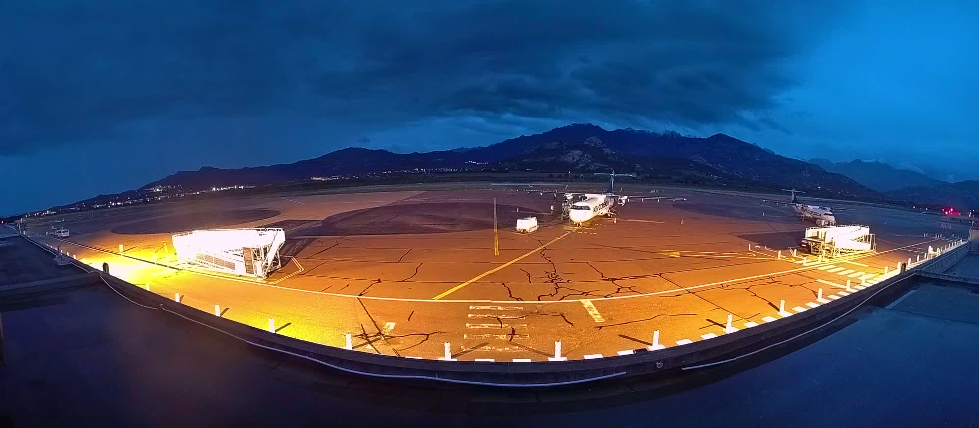 An airplane is parked on a tarmac at night, illuminated by yellow lights, with mountains in the background under a dark, cloudy sky.